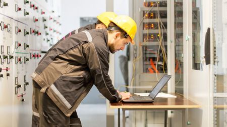 A technician in a hard hat and work jacket uses a laptop at a workstation in front of a wall of electrical controls.