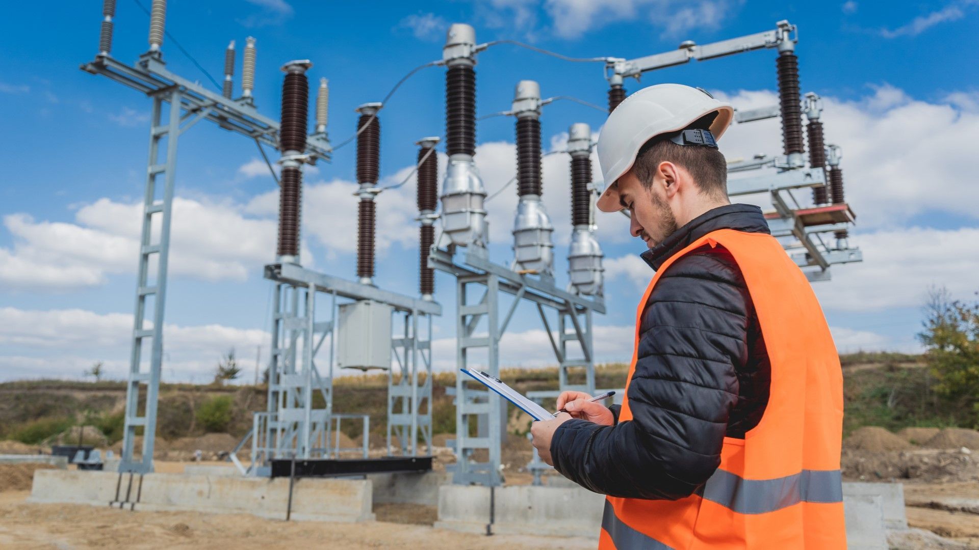 Electrician in orange vest and hardhat inspecting a power substation under a blue sky.
