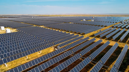 An aerial view of a vast solar farm featuring rows of dark photovoltaic panels arranged across a dry, arid landscape.