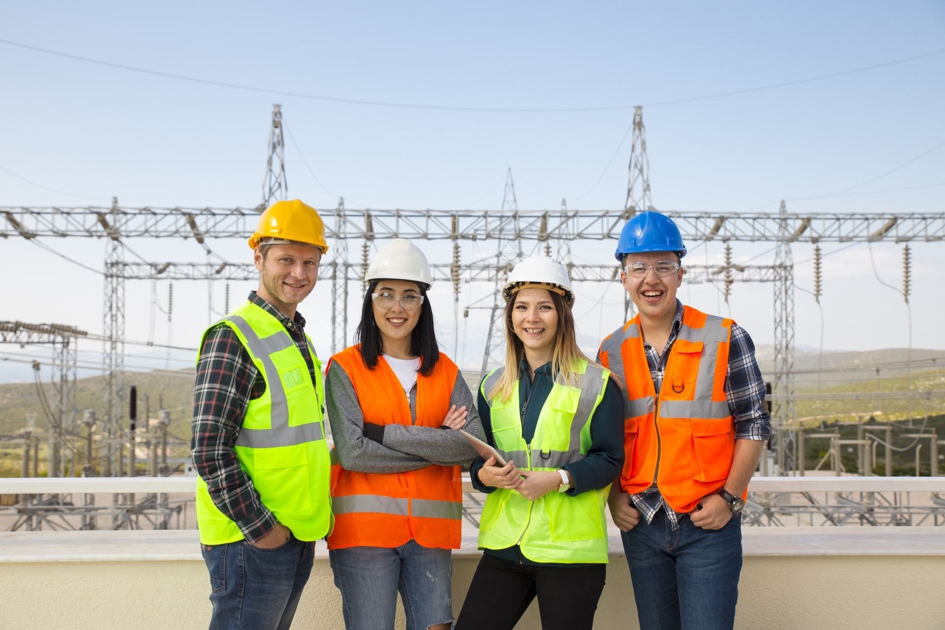 Four construction workers in safety vests and hard hats pose in front of a power station.