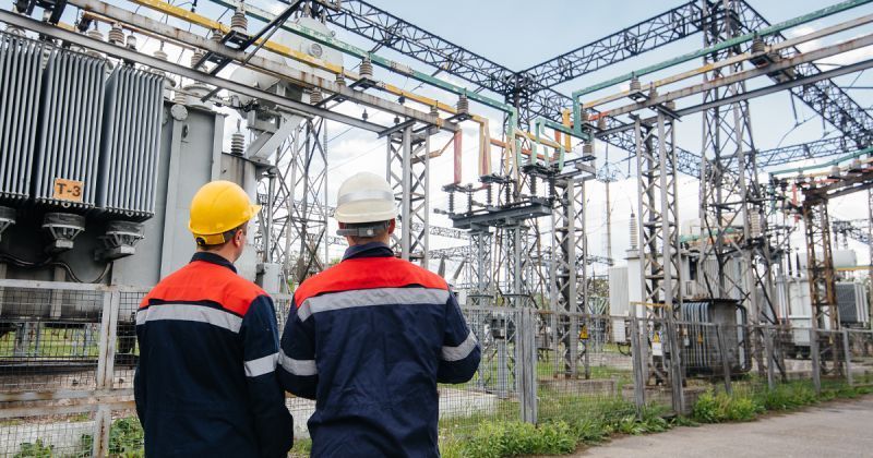 Two workers in safety vests and helmets inspect an electrical substation.
