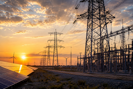 A person in a hard hat and high-visibility vest uses a tablet in front of a metal transmission tower against a blue sky.