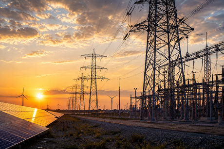 A person in a hard hat and high-visibility vest uses a tablet in front of a metal transmission tower against a blue sky.