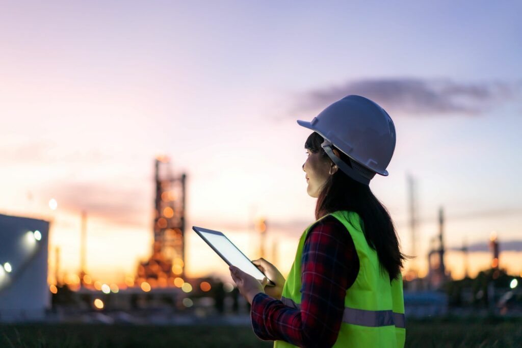 Woman in a hard hat and safety vest, holding a tablet, looking toward a factory at sunset.