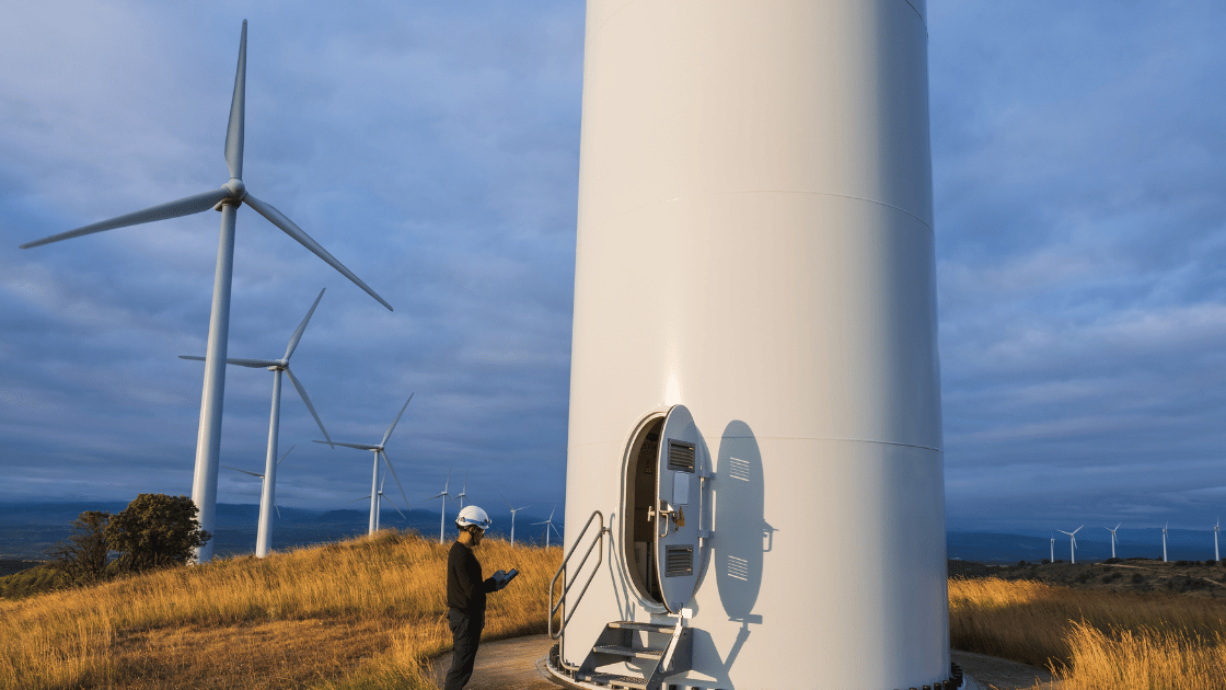 Wind turbine technician at the base of a white turbine, other turbines in background on a grassy hill, under a cloudy sky.