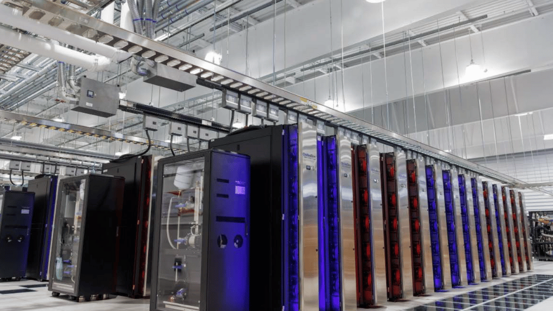 Rows of servers in a data center; silver, blue, and black cabinets with hanging cables in a bright, industrial space.