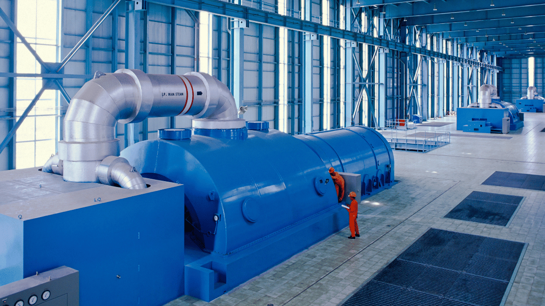 Blue industrial turbine inside a large building with a worker in orange overalls.