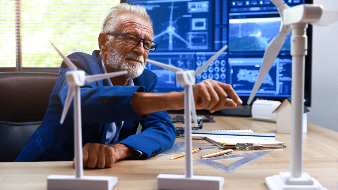 Older man in glasses points at wind turbine model, with blueprints and computer screens in background.