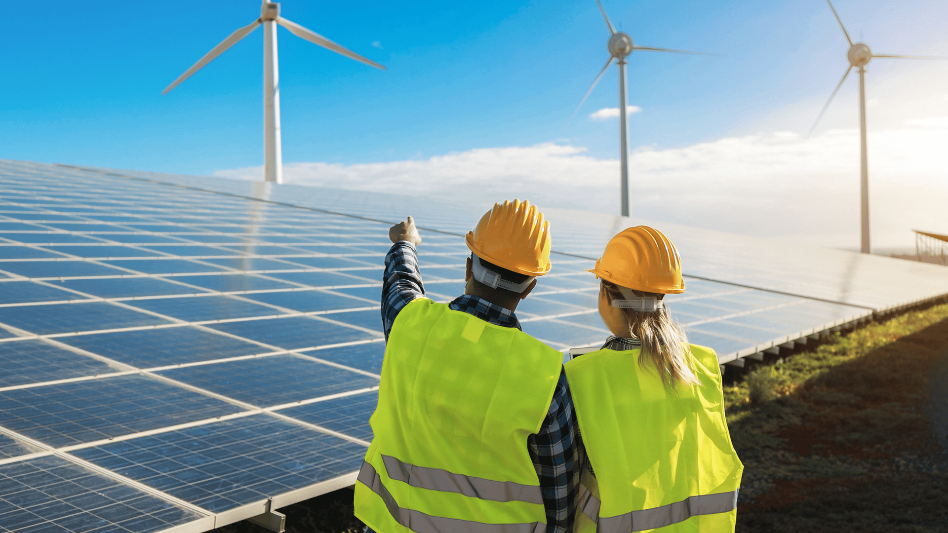 Two people in safety vests and helmets pointing at wind turbines and solar panels.