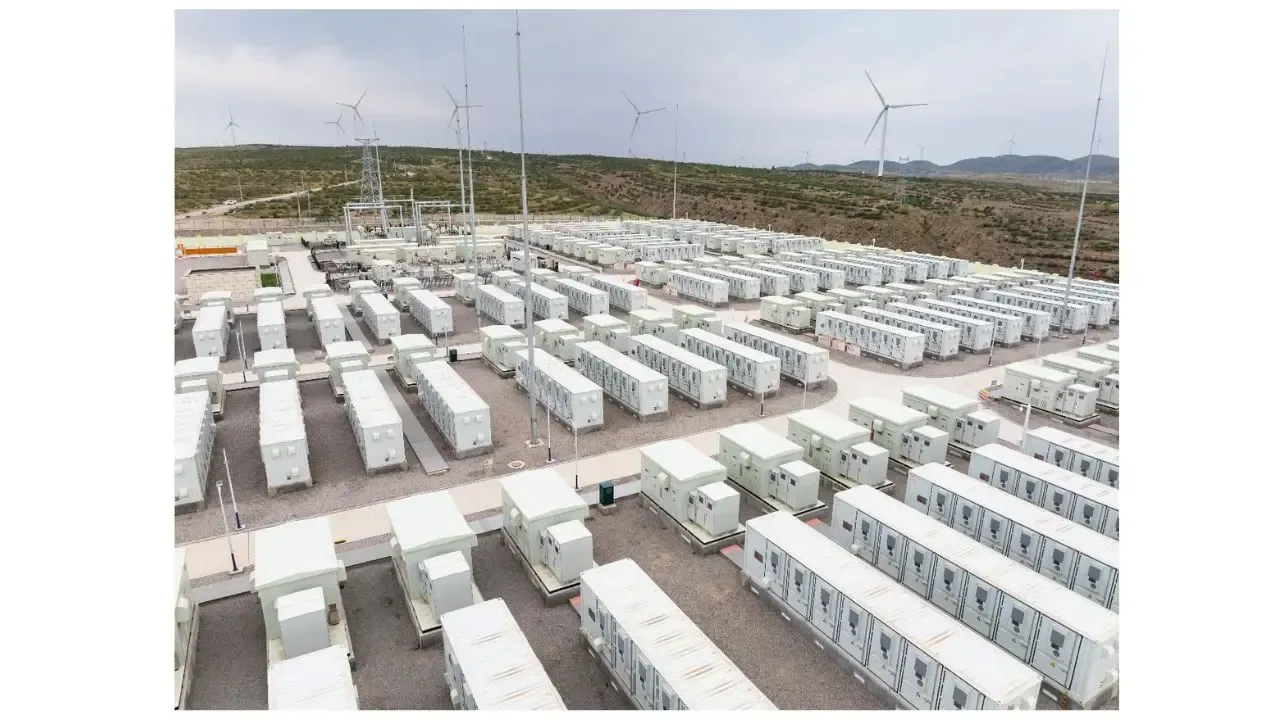 An aerial view of a large battery storage facility with numerous white shipping container-like units. Wind turbines in background.