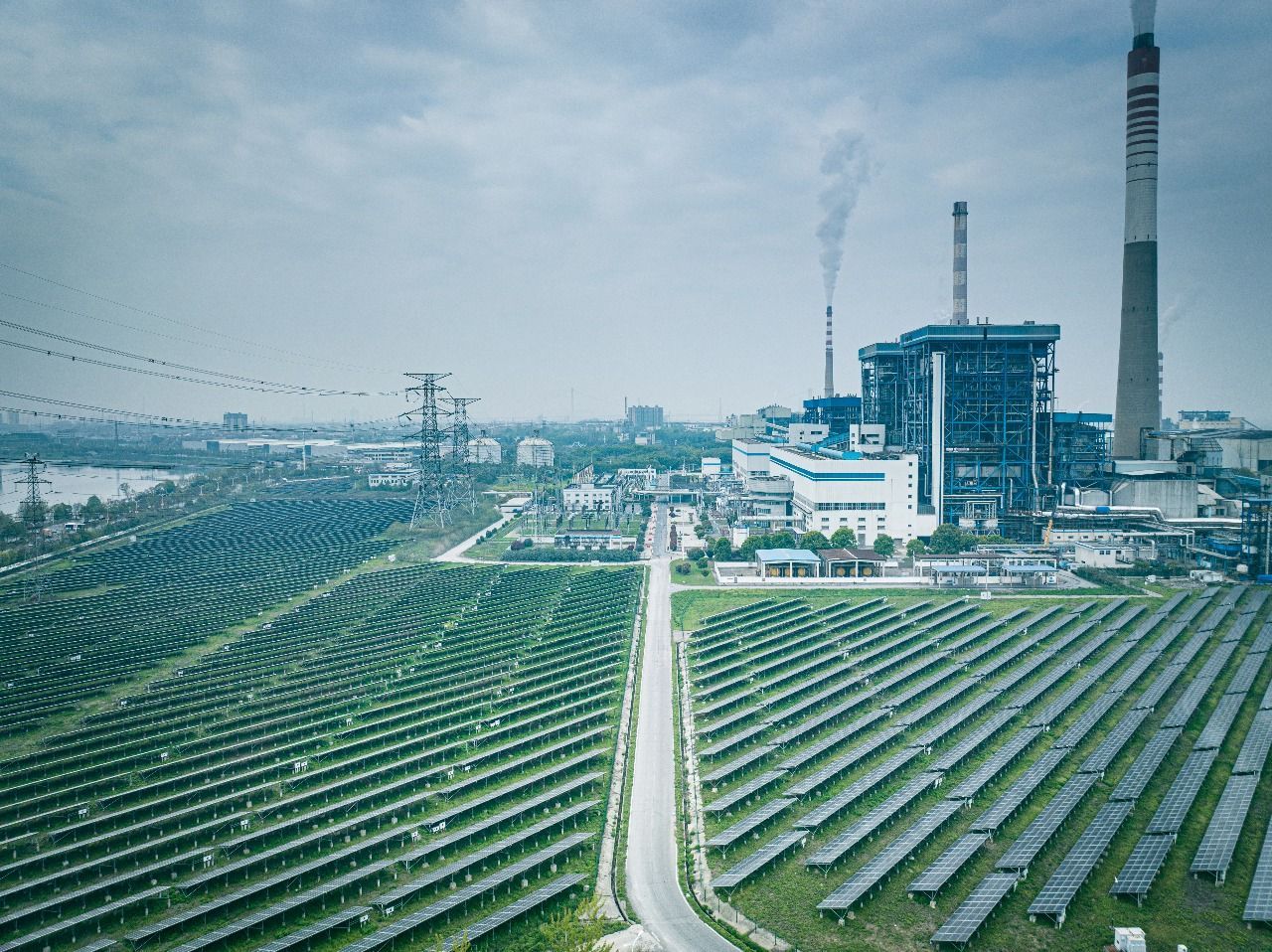 Aerial view of rows of solar panels extending toward a large industrial power plant with smoking chimneys.