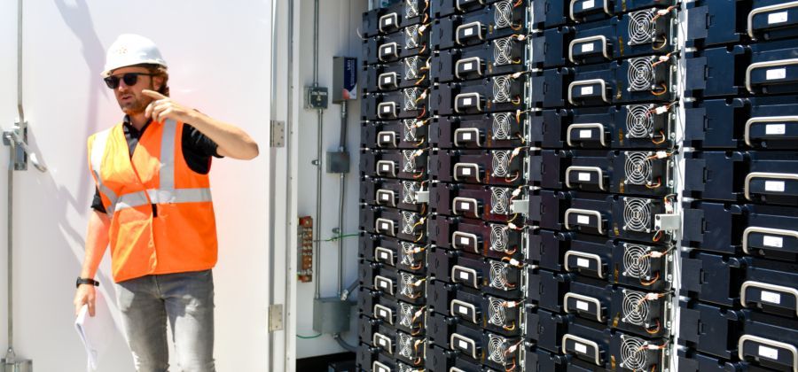 Man in safety vest and hard hat points to a wall of black battery modules.