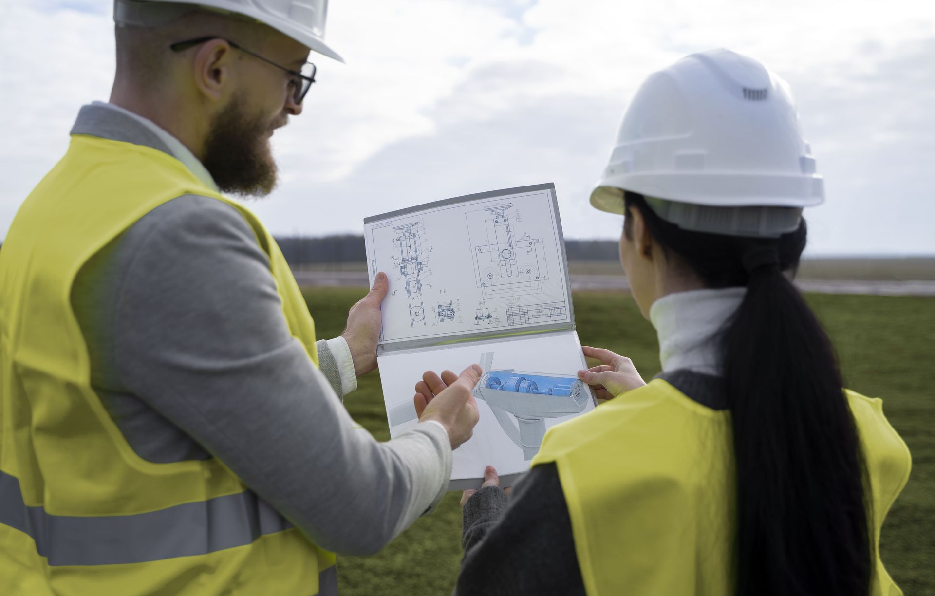 Two people in hard hats and vests review blueprints outdoors.