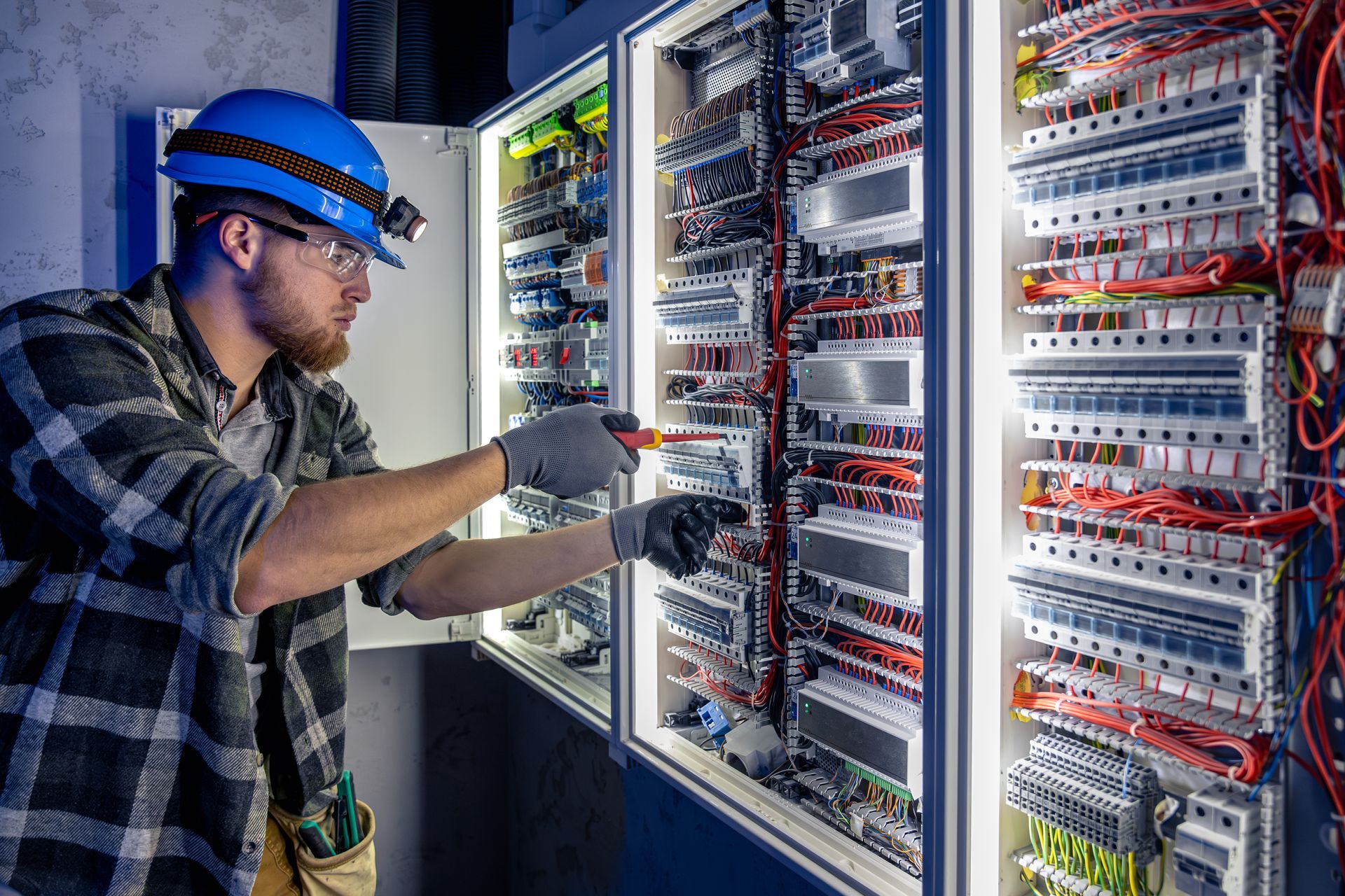 Electrician in blue hardhat and safety glasses, working on a complex electrical panel.