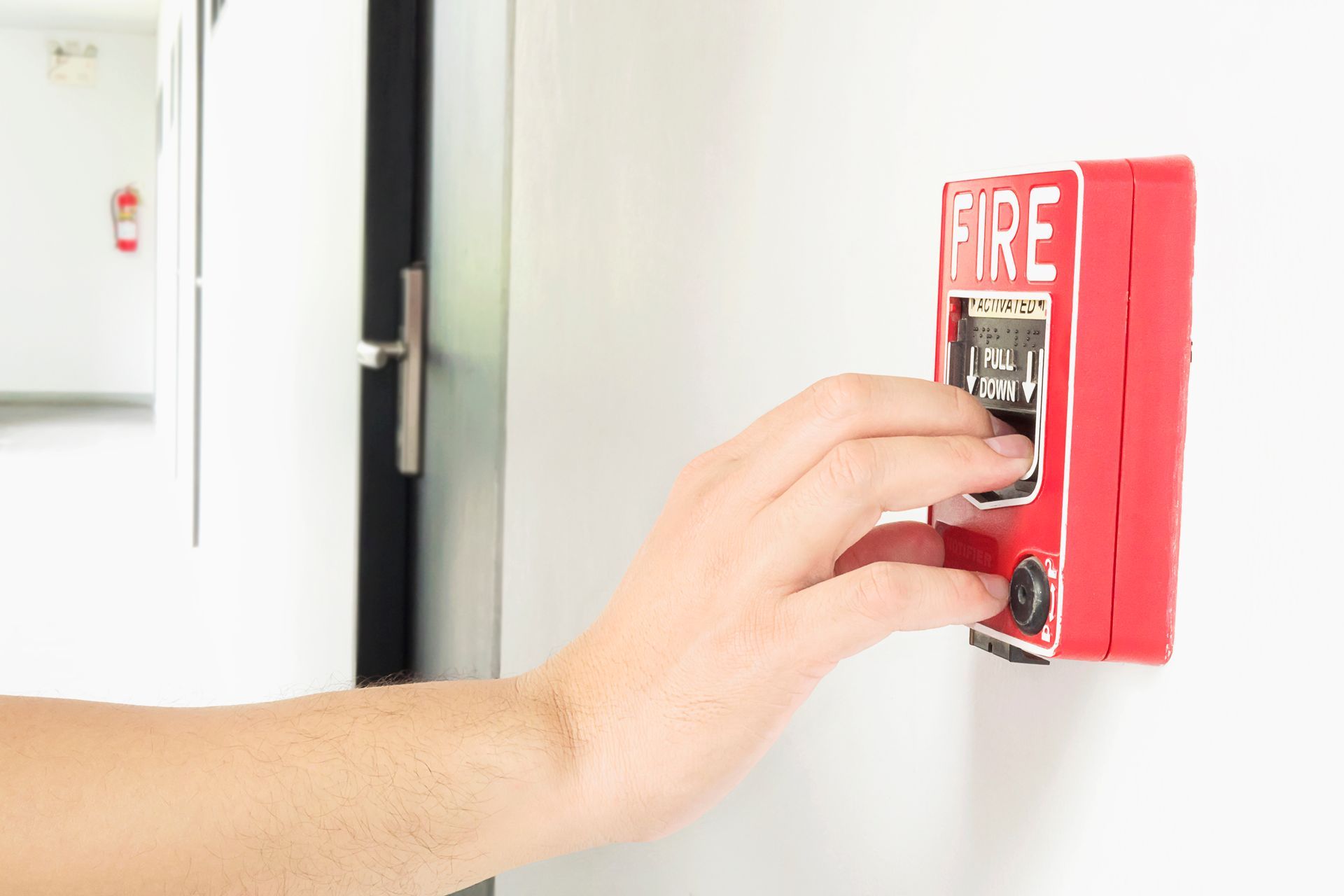 Hand activating a red fire alarm on a white wall in a hallway.