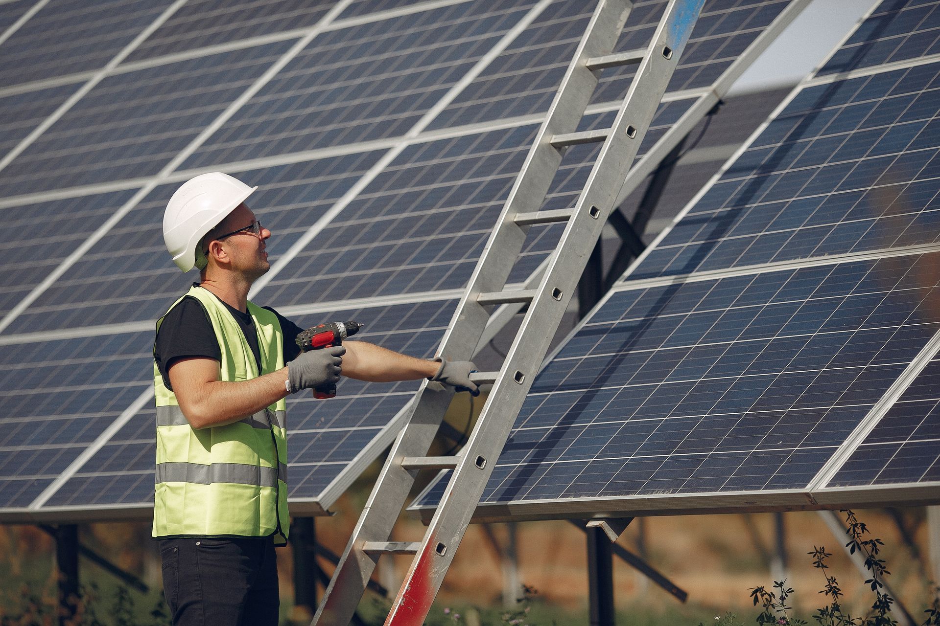 Man in hard hat and safety vest using a drill on solar panels, standing on a ladder.