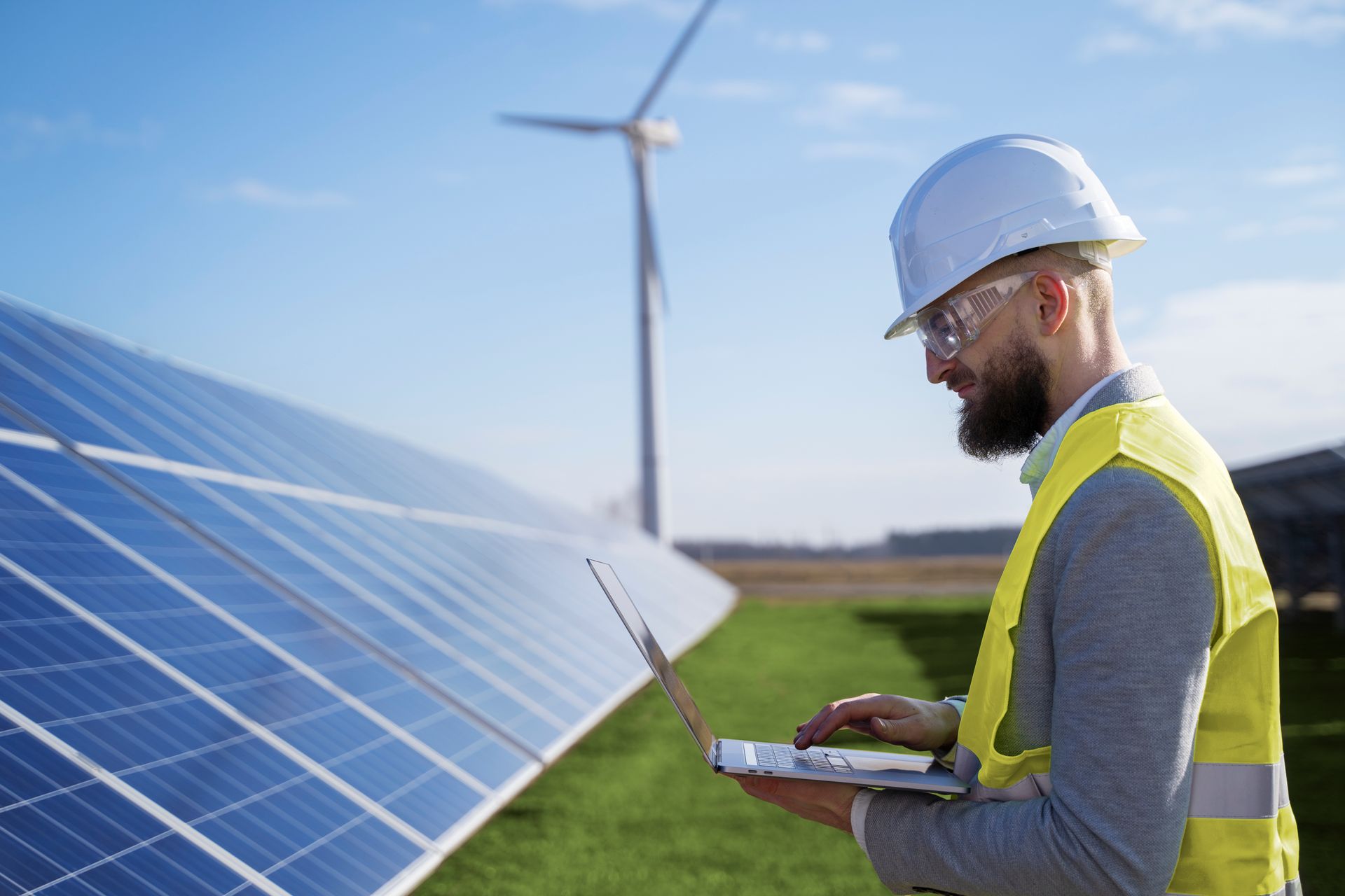 Engineer wearing safety gear uses a laptop near solar panels and a wind turbine.
