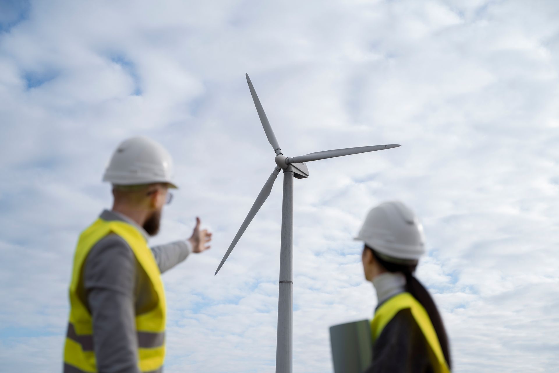 Two engineers in hard hats and vests inspecting a wind turbine against a cloudy sky.