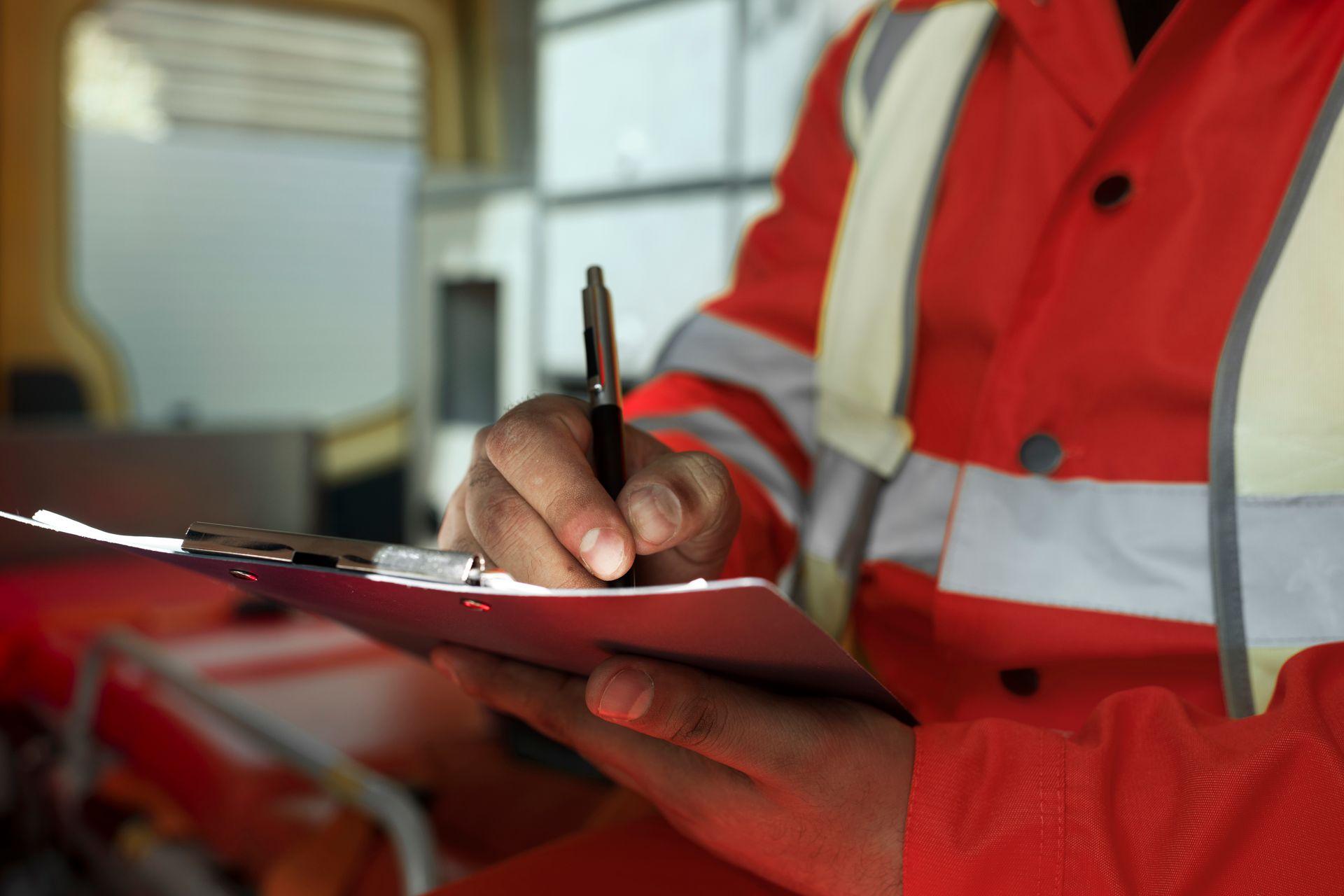 Person in an orange vest writing on a clipboard, possibly an ambulance, indoors.