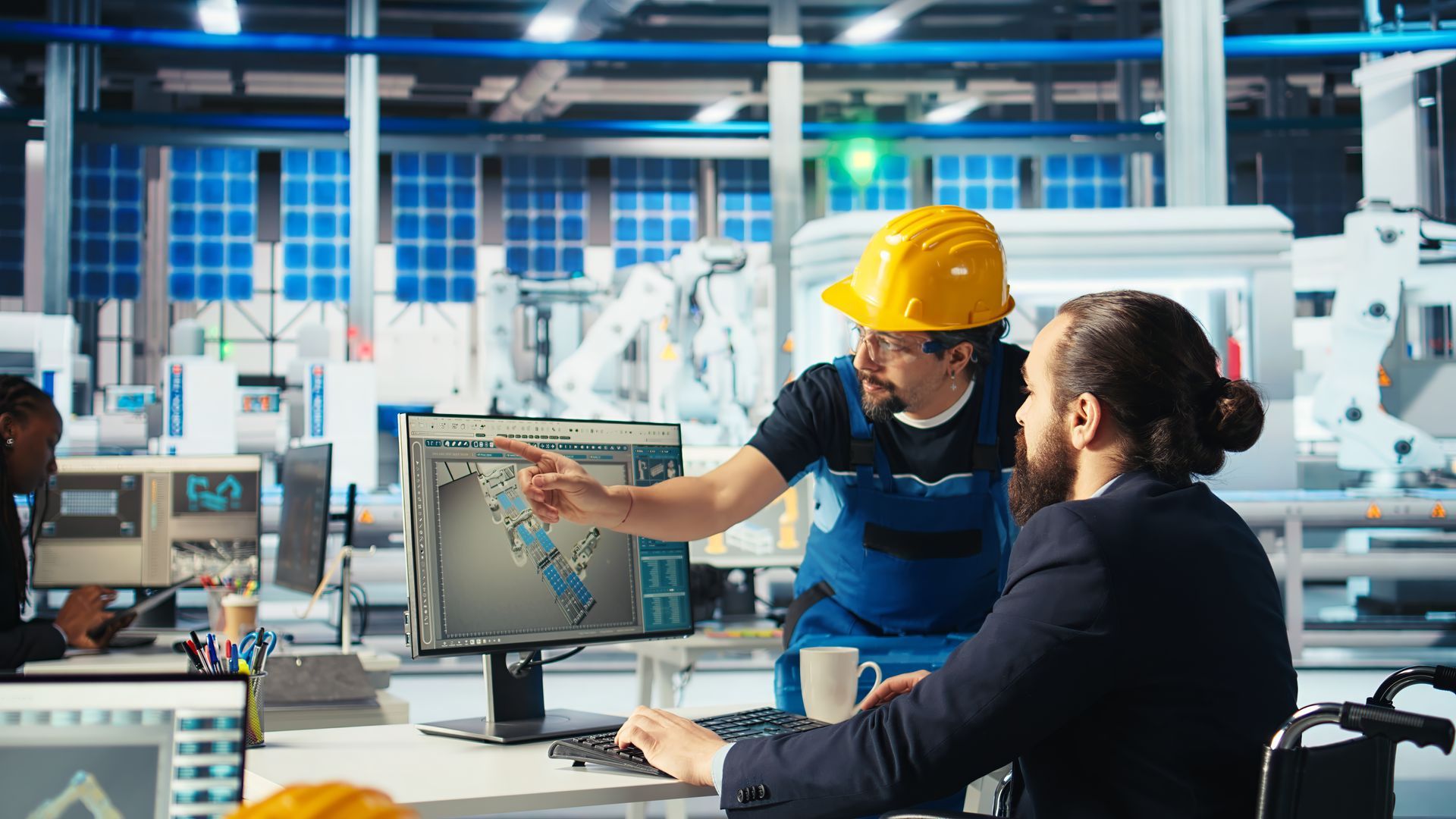 Man in hard hat points at a computer screen to a person in a suit, factory setting.