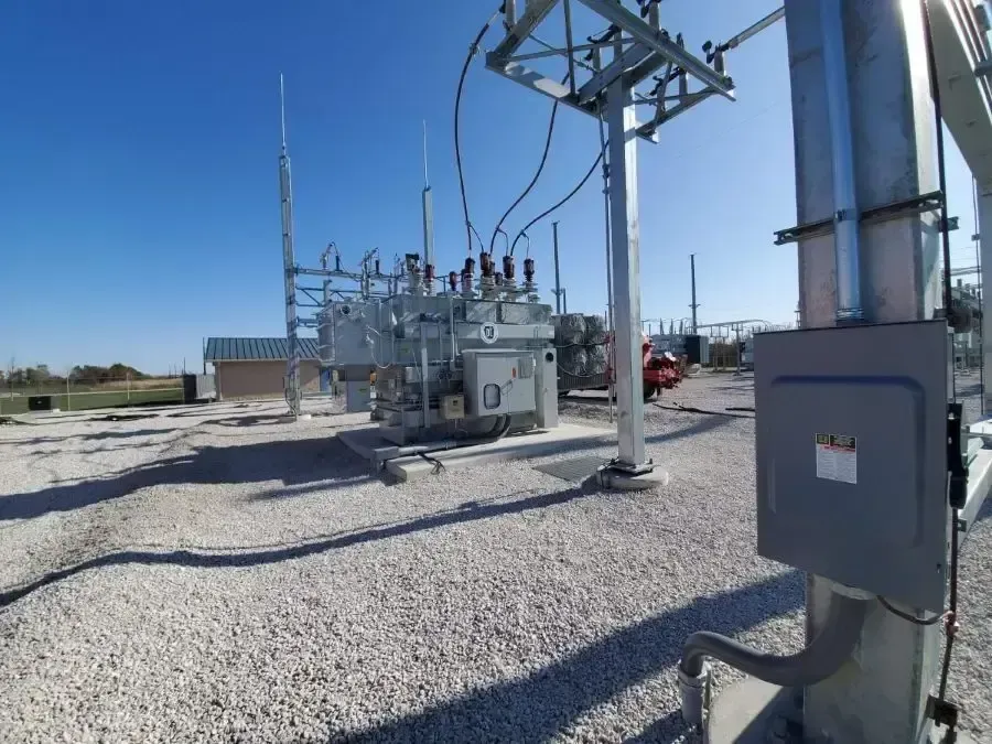 Electrical substation with transformers and equipment against a clear blue sky. Gravel ground and metal structures.