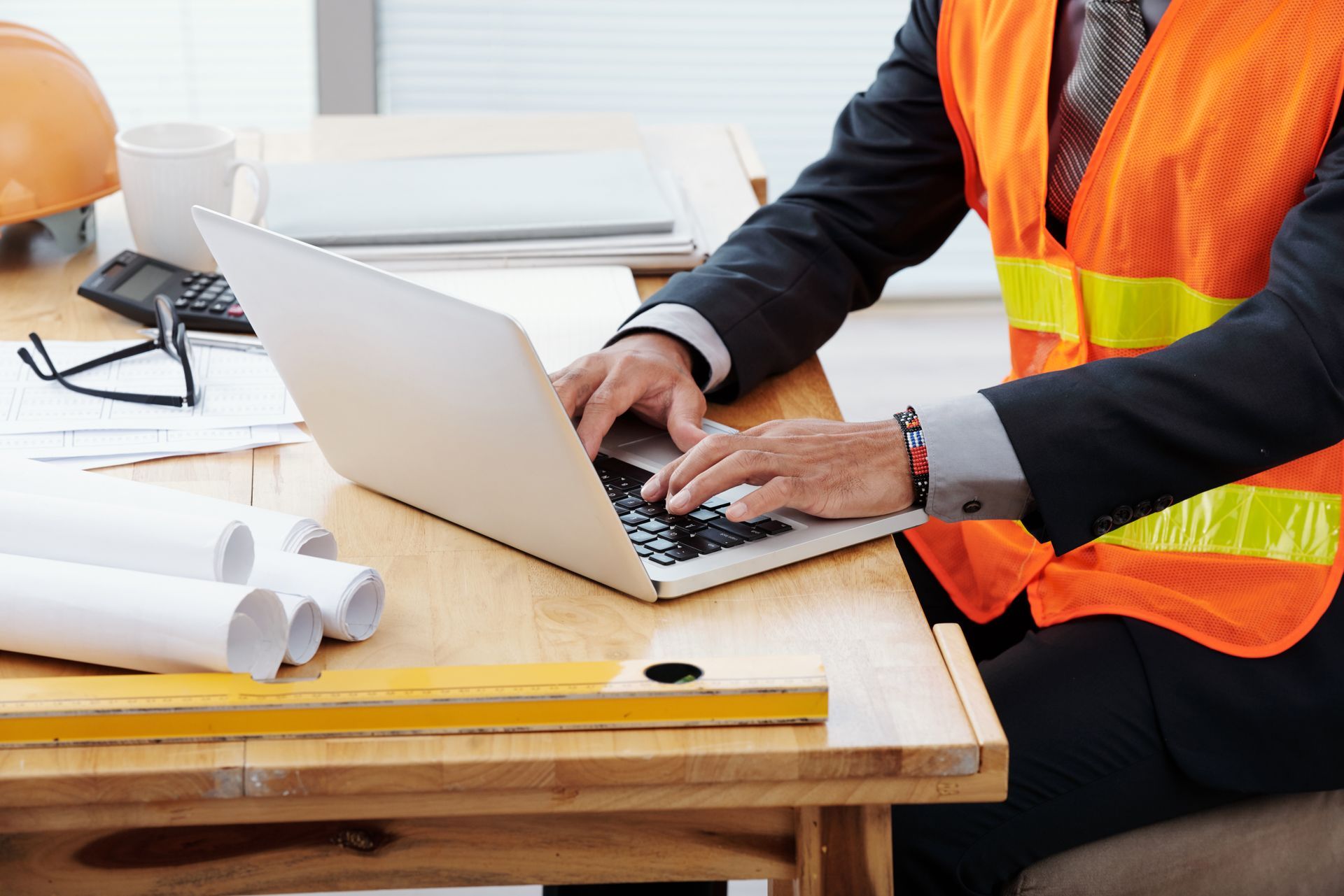 Person in construction vest typing on a laptop at a desk with blueprints, a level, and a hard hat.