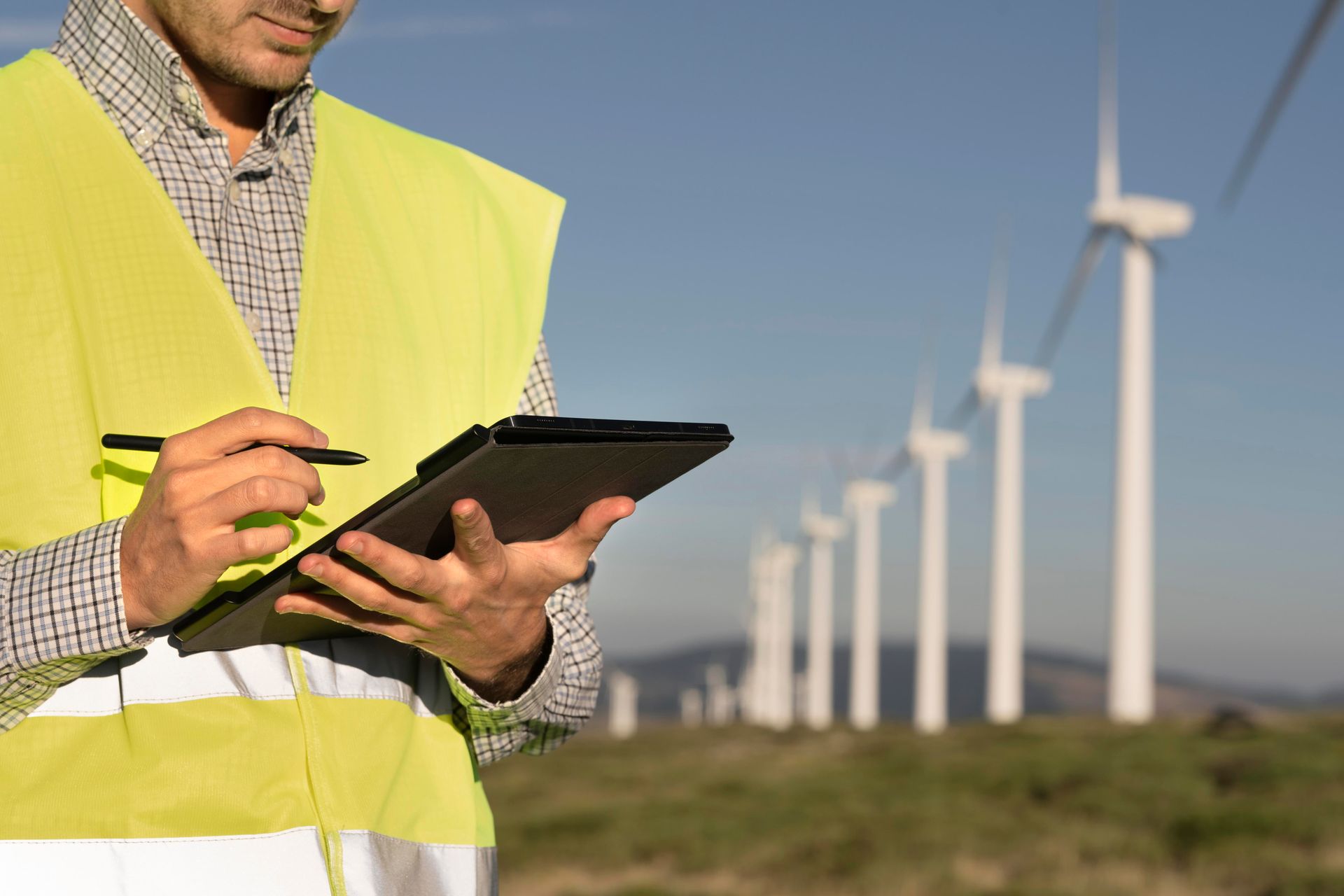 Engineer in reflective vest holding tablet, inspecting wind turbines in a field.