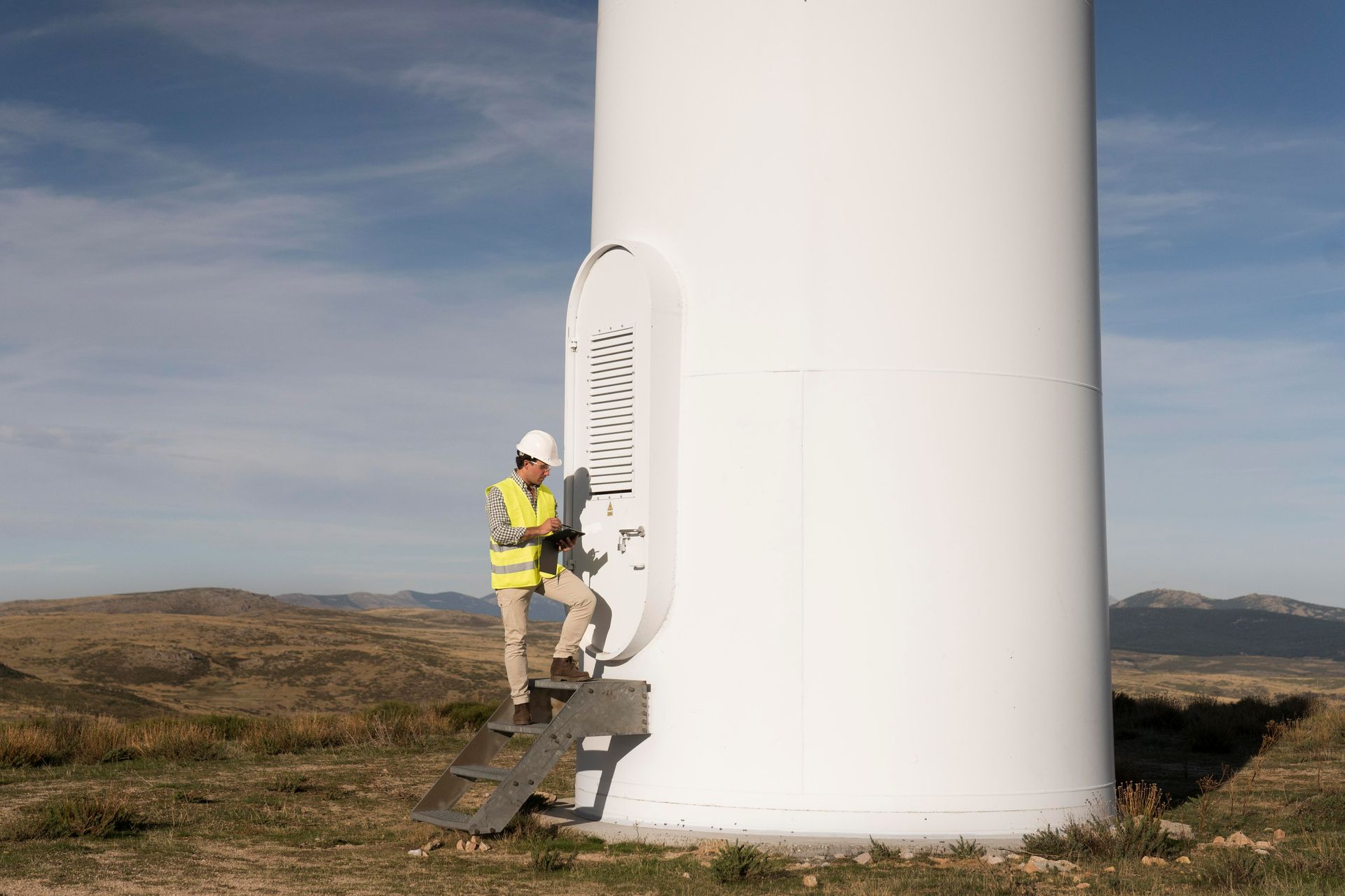 Person in safety gear climbing steps on a white wind turbine in a field.
