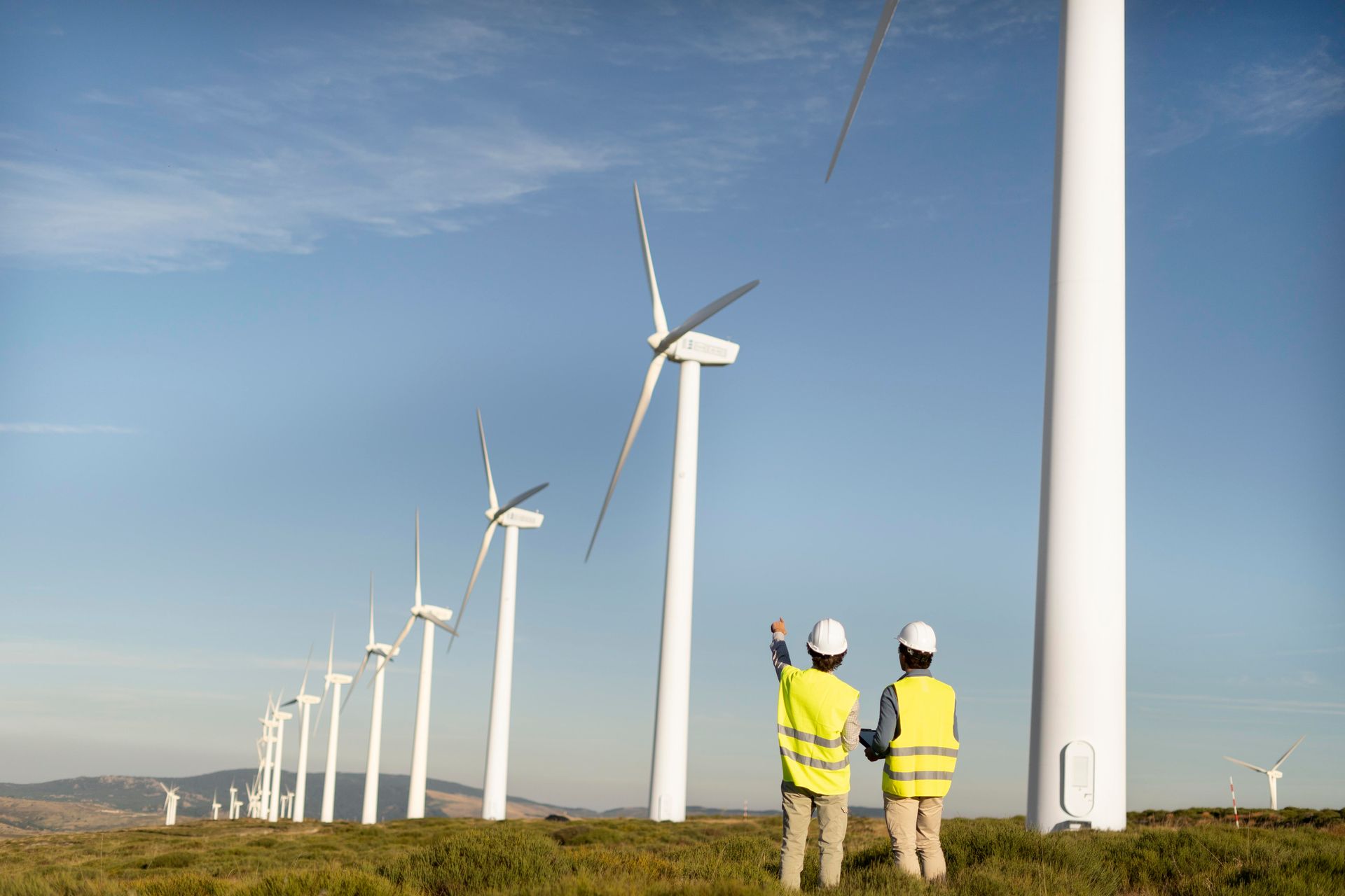 Two people in safety vests at a wind farm, pointing toward the turbines, on a sunny day.