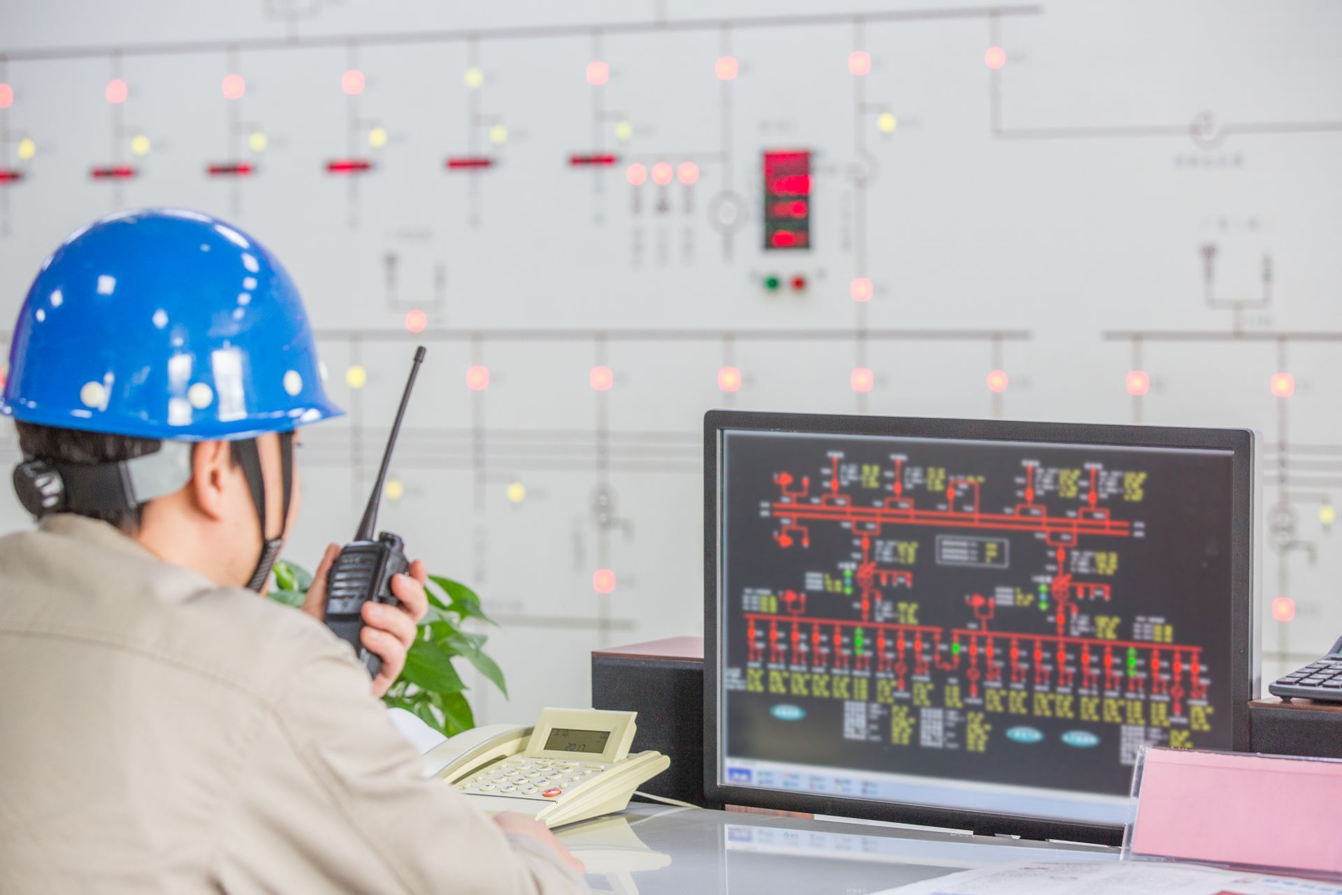 Person in blue hard hat uses a radio in a control room, looking at a computer screen displaying electrical schematics.