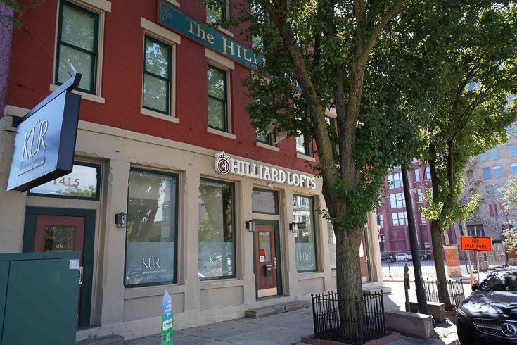 Street view of a red brick storefront with a tree and parked cars on a sunny city block