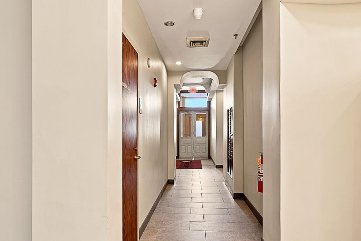 Narrow apartment hallway with white walls, wood doors, tiled floor, and a bright doorway at the end