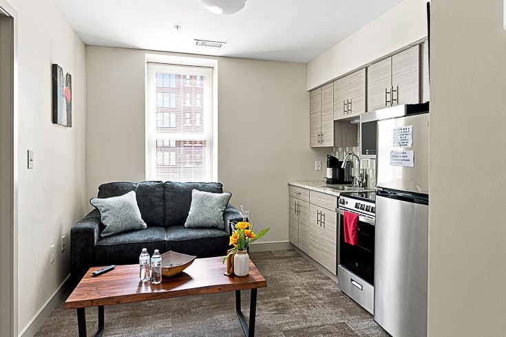 Small modern kitchen-living room with gray sofa, wood coffee table, and white cabinets.