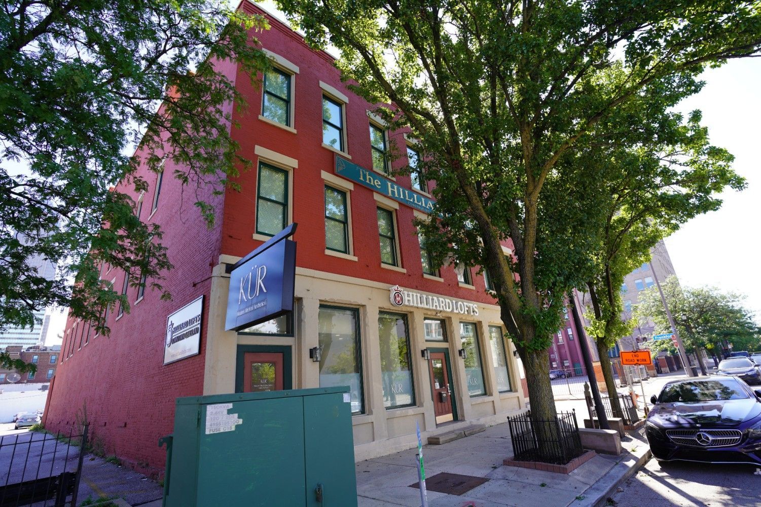 Red-brick corner storefront with large windows on a tree-lined street