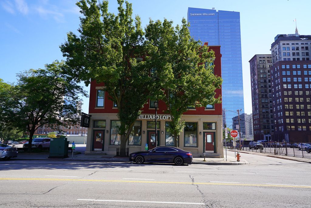 Small storefront under leafy trees on a city street, with a parked car and tall buildings in the background.