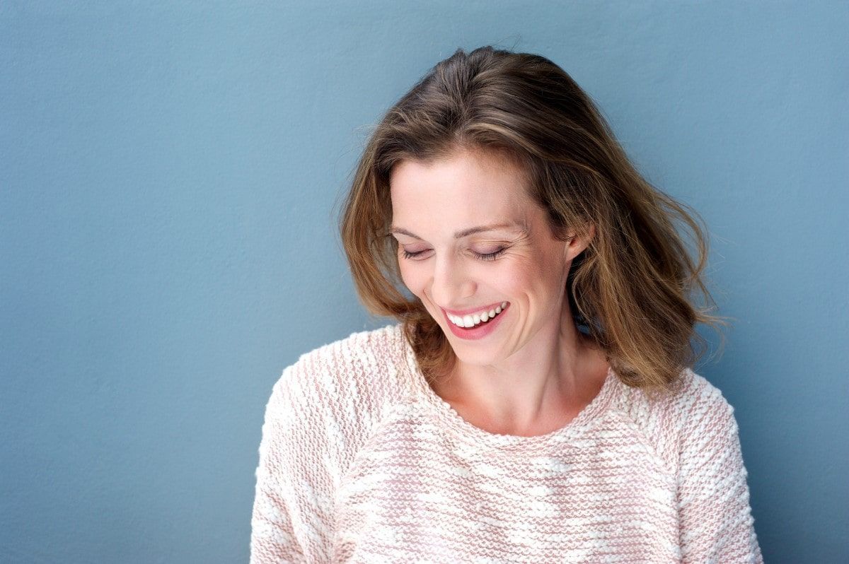 Woman with brown hair smiling, looking down, wearing a white sweater against a blue background.