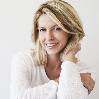 Blonde woman smiles, resting chin on hand, wearing white sweater, against white background.