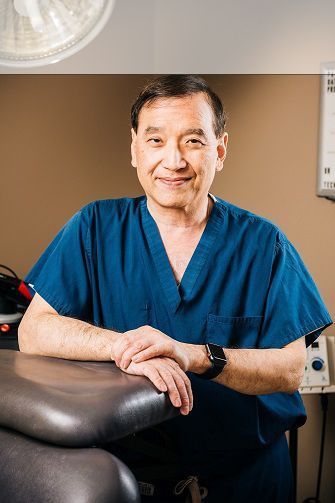 Doctor in blue scrubs smiling, leaning on a medical table, in an exam room.