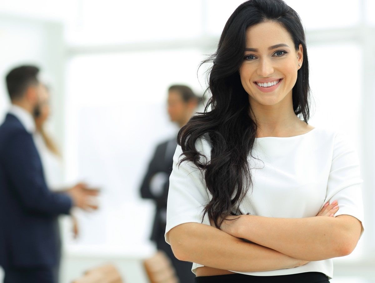 Woman with dark hair, white blouse, arms crossed, smiling in office.