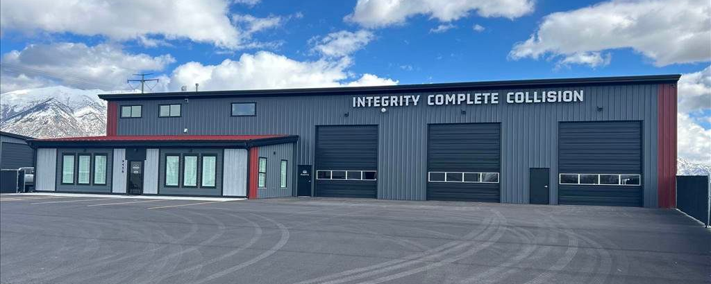 Exterior of a metal building with garage doors, office windows, and a mountain backdrop under a cloudy sky.