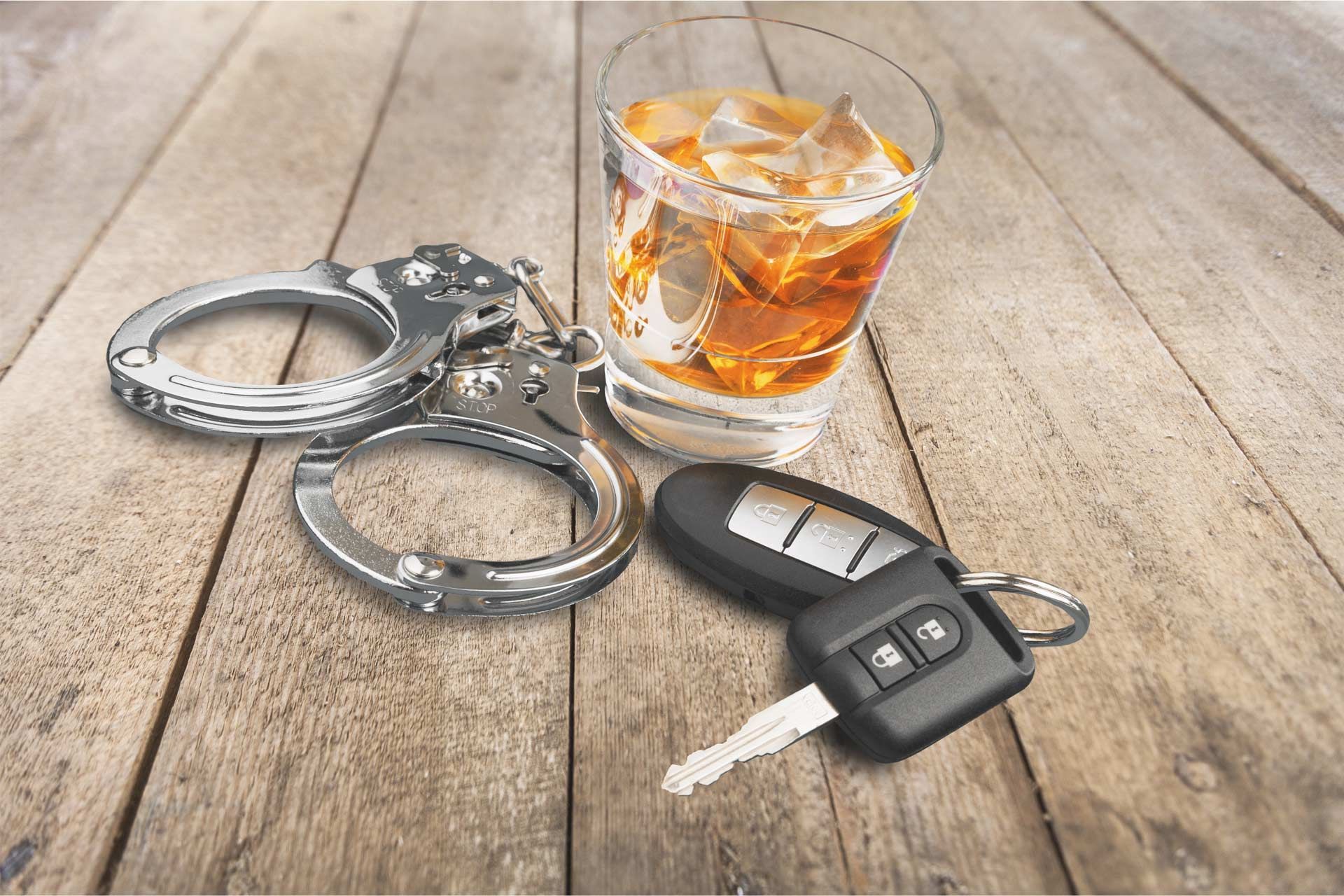 A glass of whiskey , handcuffs , and a car key on a wooden table