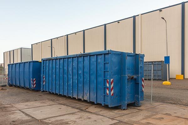 A row of blue dumpsters are parked in front of a building. A row of blue dumpsters are parked in front of a building.