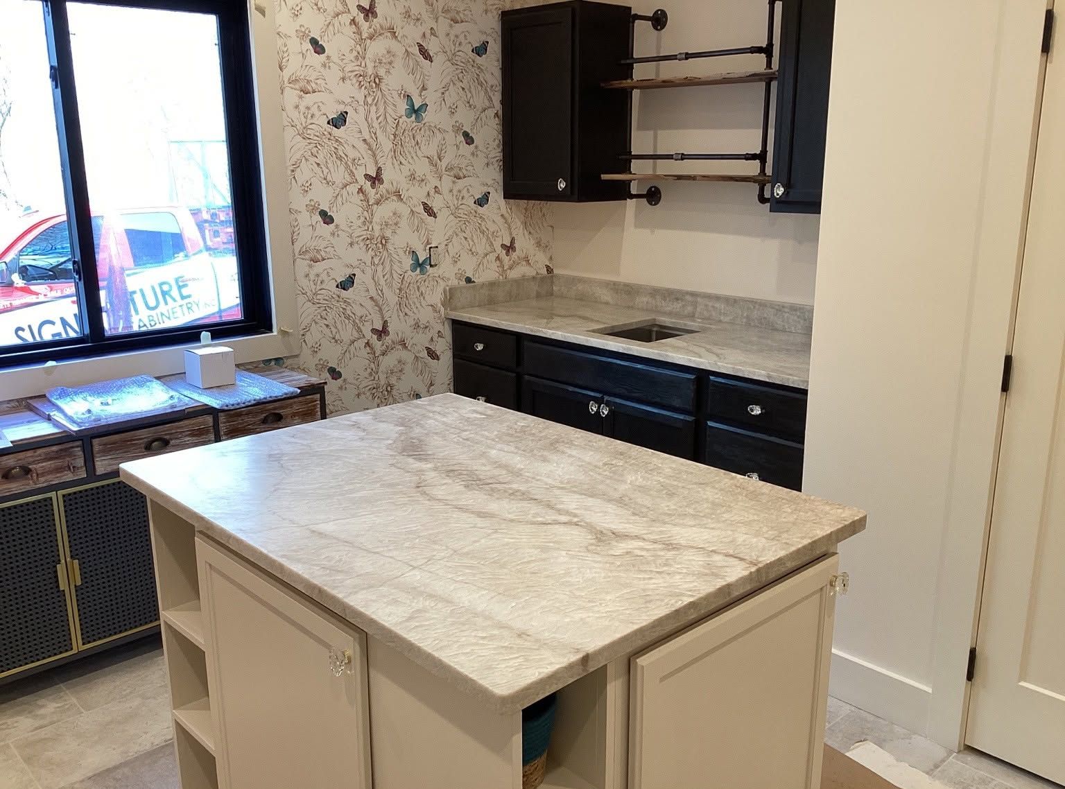 Empty kitchen with white island, dark cabinets, patterned wallpaper, and a window by the sink.