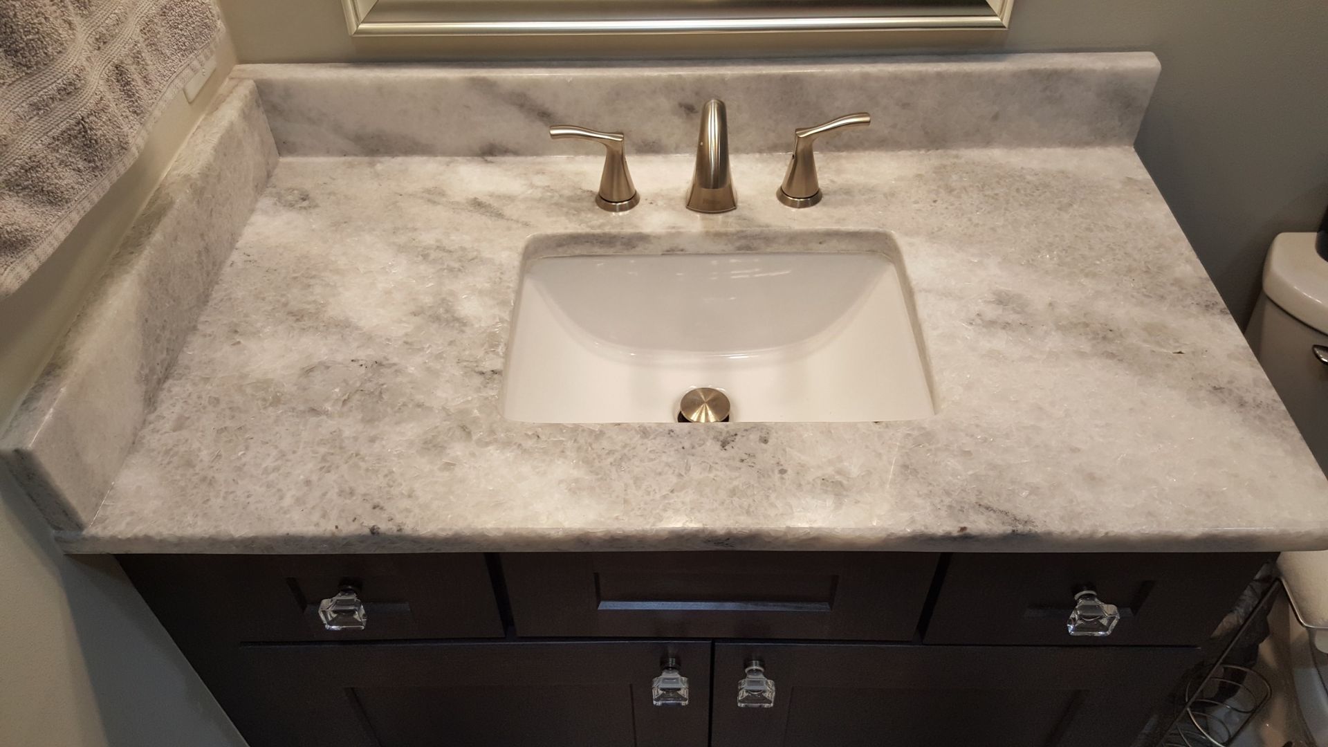 Bathroom sink with light stone countertop, bronze faucet, and dark wood vanity below mirror