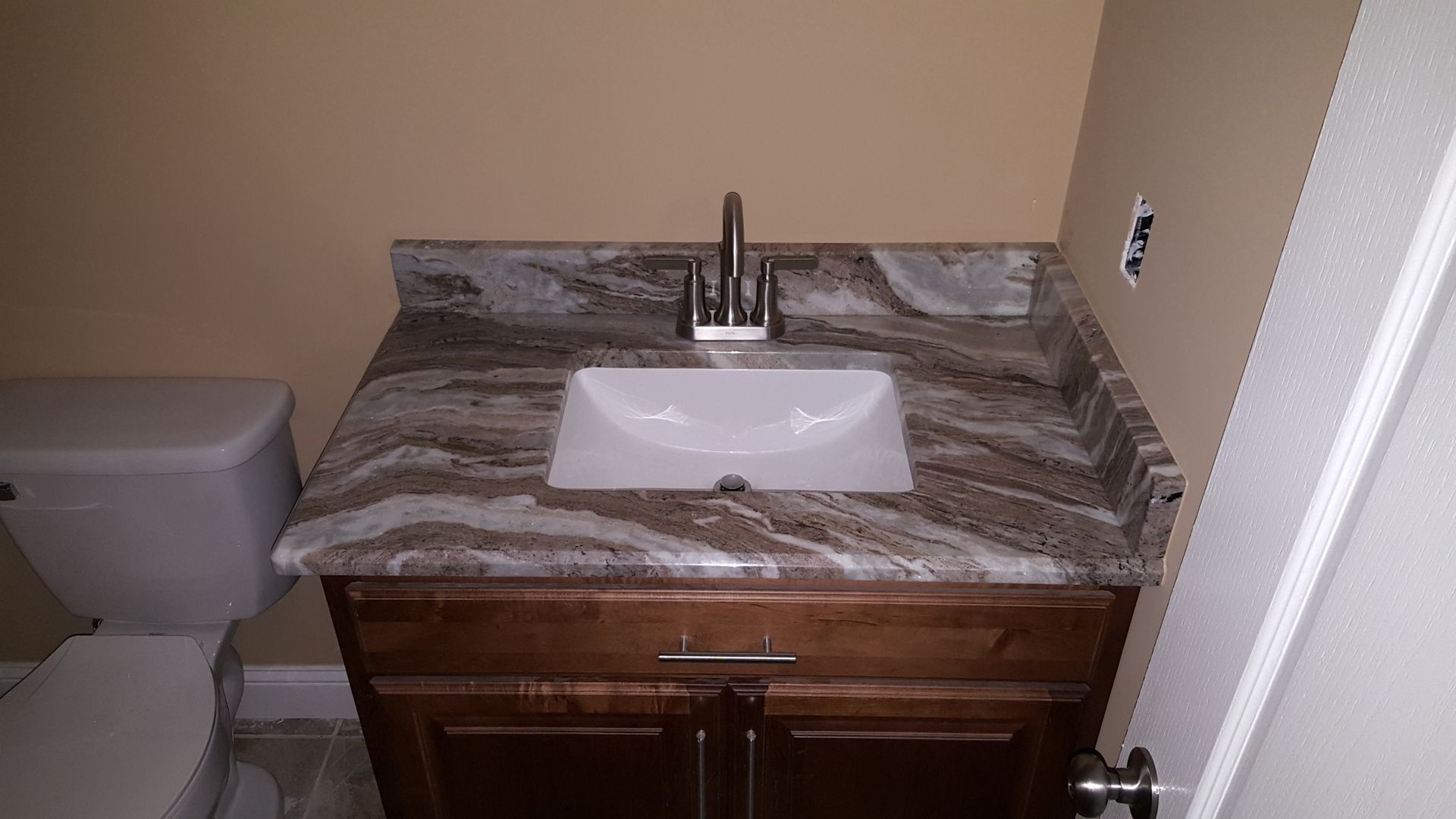 Bathroom vanity with white sink, brown cabinet, marble countertop, and toilet on the left.