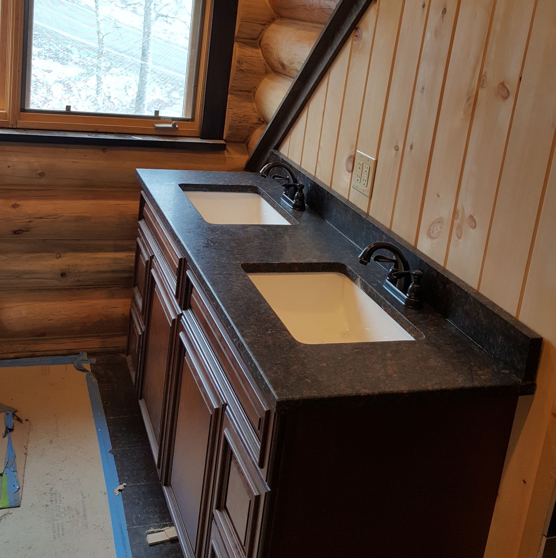 Wooden bathroom vanity with double sinks, black countertop, and window in a rustic room