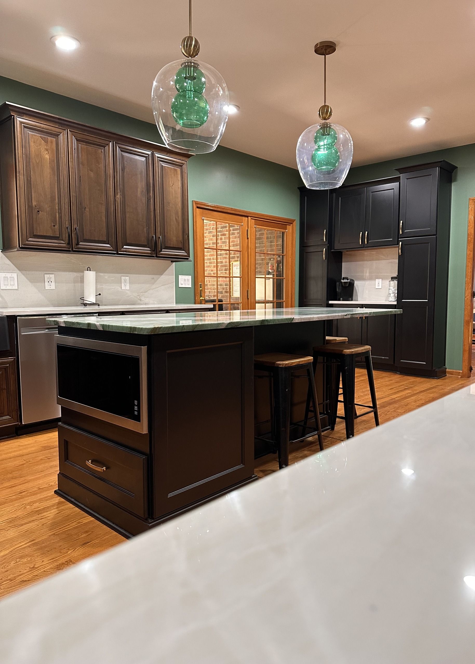 Modern kitchen with dark cabinets, island stools, pendant lights, and a window with wooden blinds.