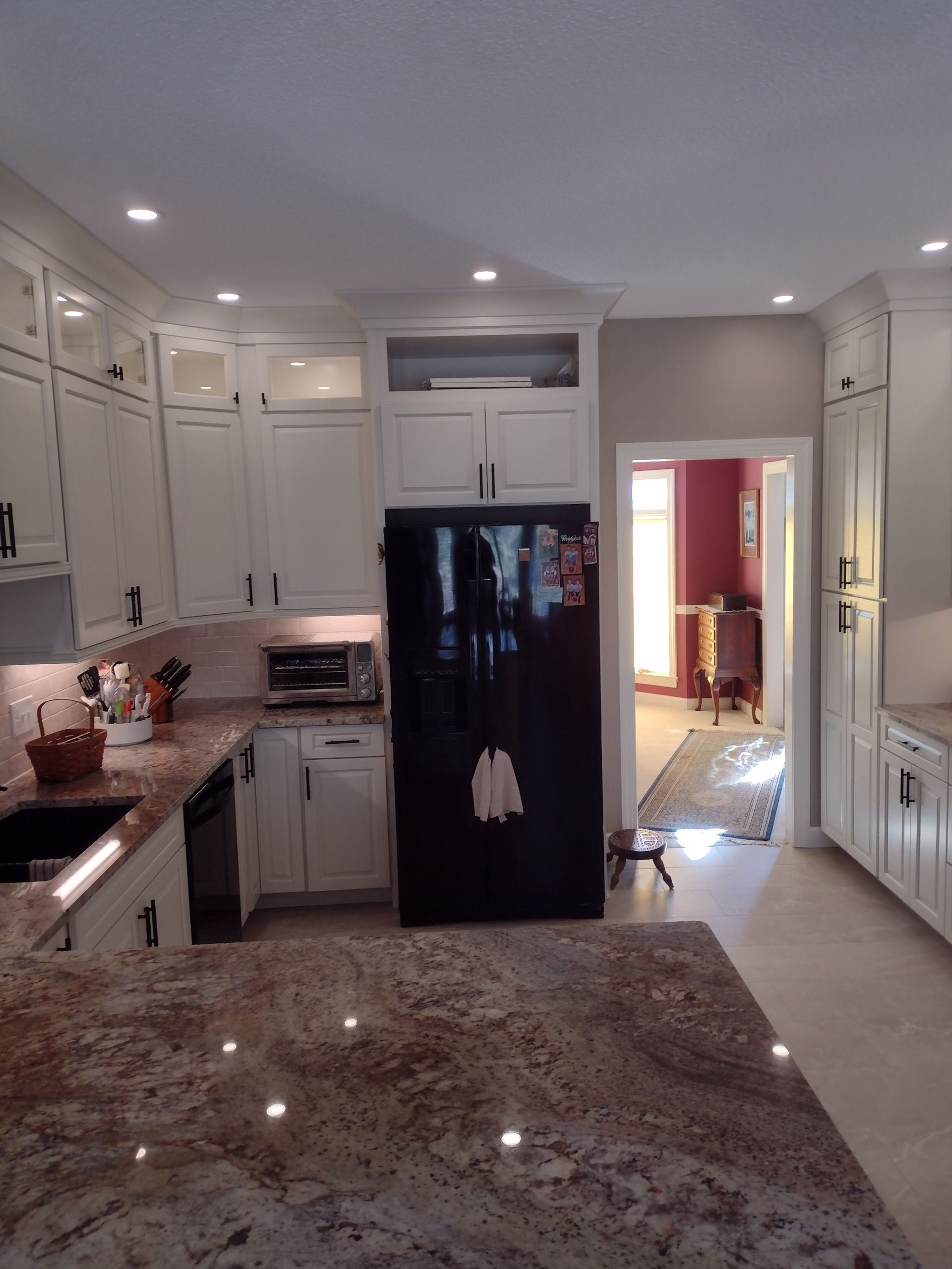 Bright kitchen with white cabinets, granite counters, central black refrigerator, and open doorway to another room