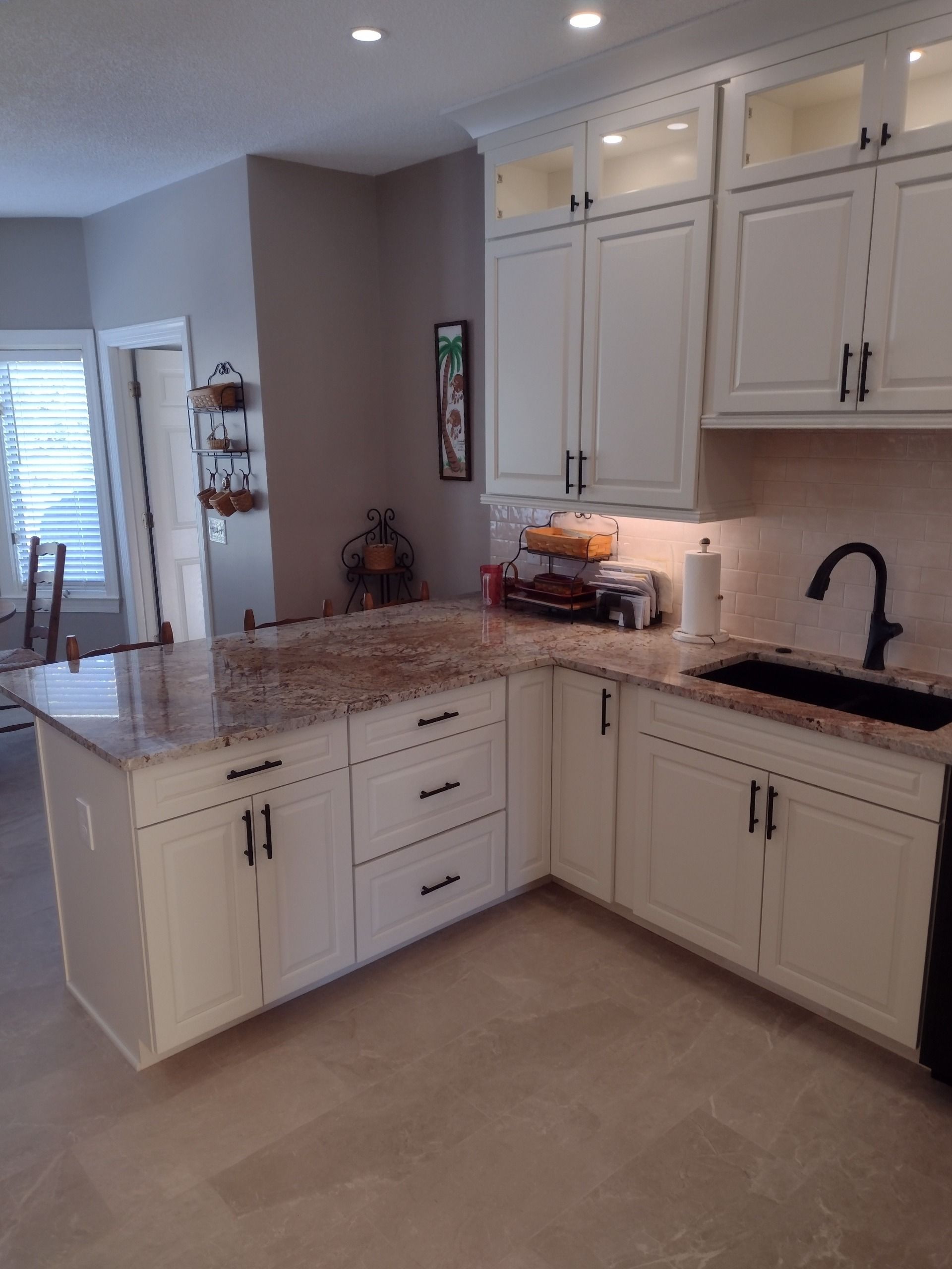 Bright kitchen with white cabinets, speckled granite island, black sink, and beige tile floor.
