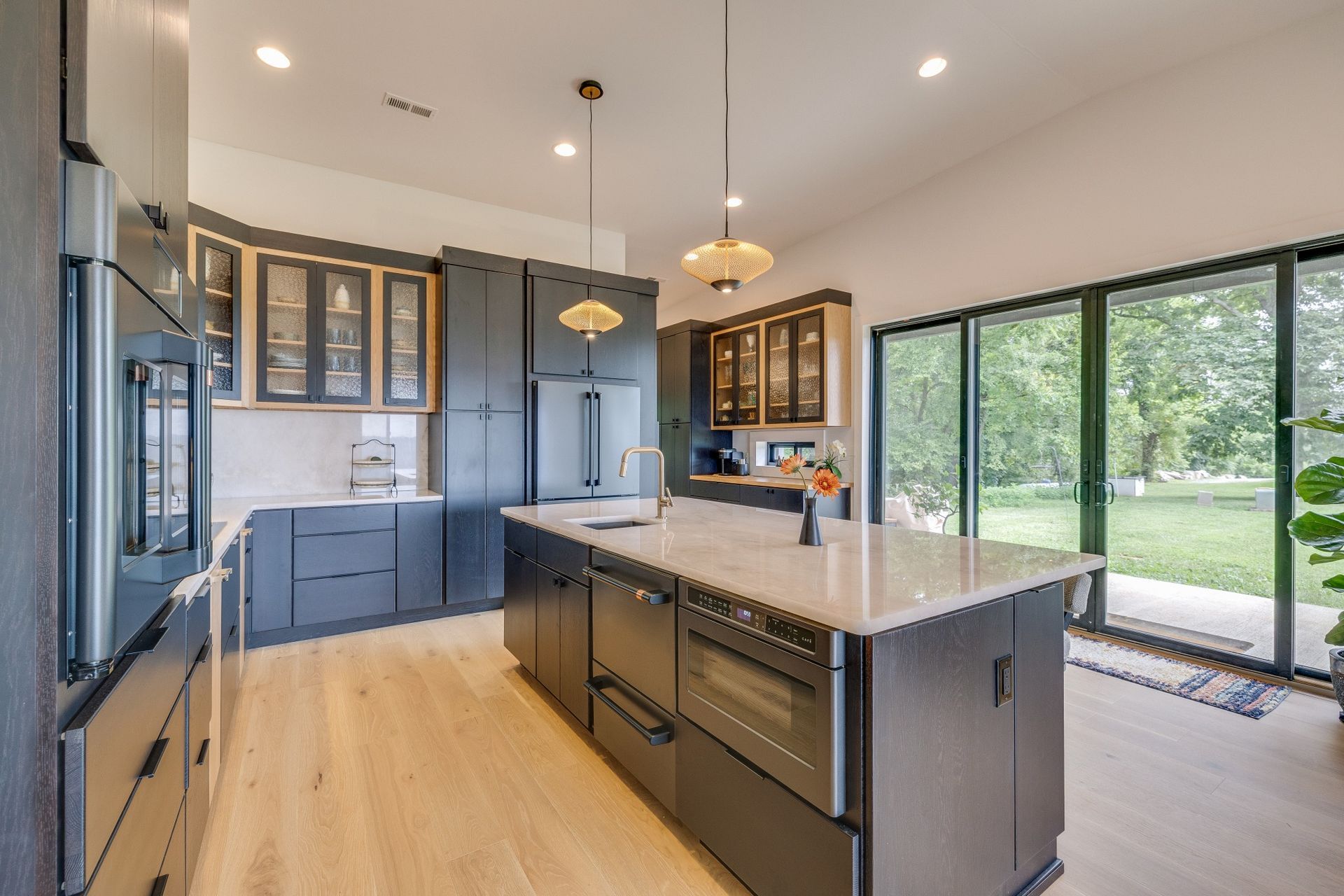 Bright modern kitchen with dark blue cabinets, large island, pendant lights, and floor-to-ceiling windows overlooking trees